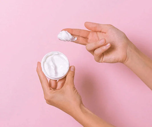 Hand holding open jar of white face cream with pink background, skincare product
