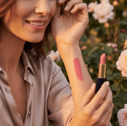 Woman applying pink lipstick and showing color swatch on arm with floral background