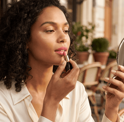 Woman applying lipstick in a casual setting with plants and furniture in the background