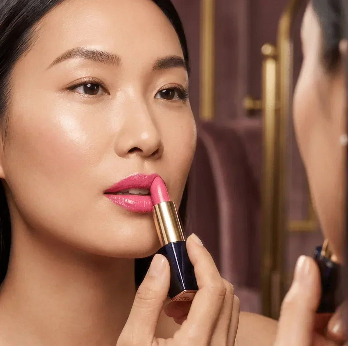 Woman applying pink lipstick with gold and navy blue tube while looking in mirror