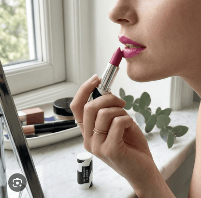 Person applying pink lipstick in front of a mirror with a blurred background